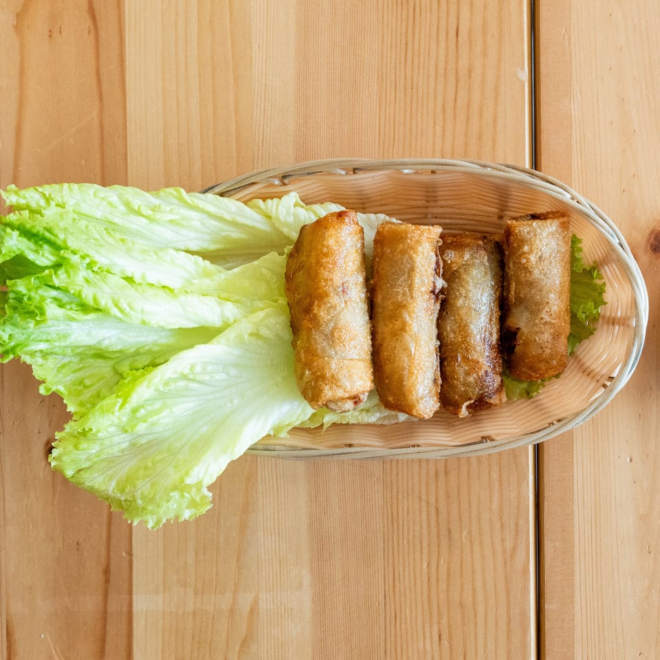 Four crispy fried spring rolls on a bed of fresh lettuce in a woven basket, with dipping sauce.