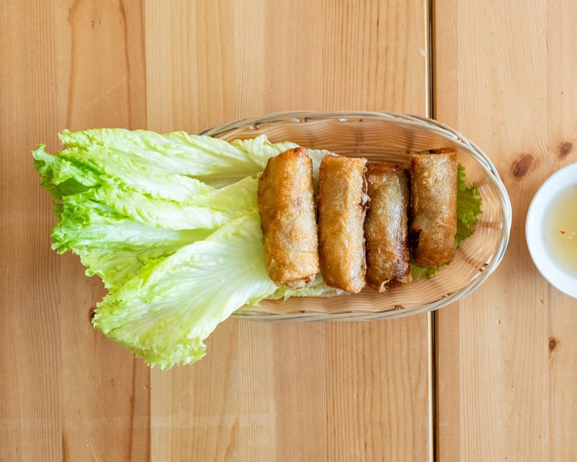 Four crispy fried spring rolls on a bed of fresh lettuce in a woven basket, with dipping sauce.