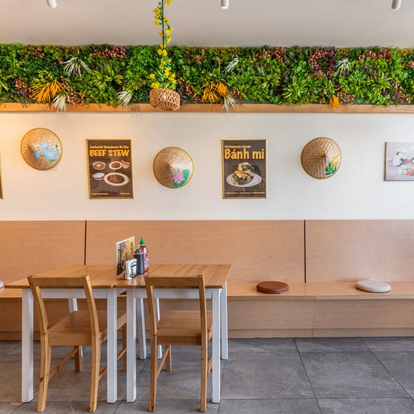 A bright Vietnamese restaurant interior with light wood tables, benches, and a green plant wall.