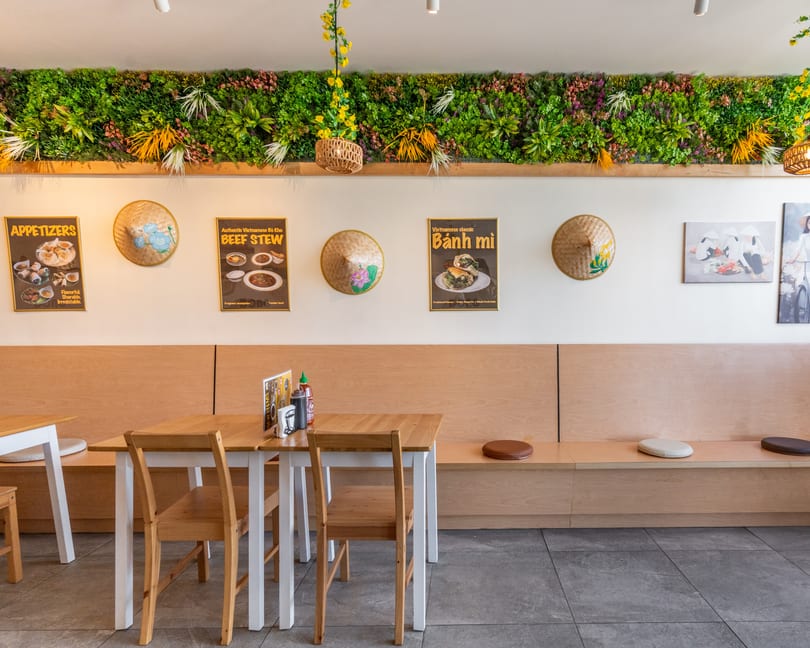 A bright Vietnamese restaurant interior with light wood tables, benches, and a green plant wall.