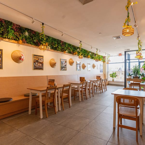 Bright, empty restaurant interior with light wood tables, chairs, and banquettes, adorned with plants and conical hats.