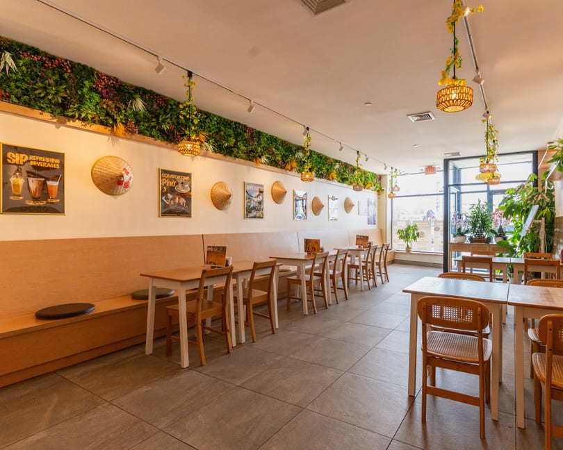 Bright, empty restaurant interior with light wood tables, chairs, and banquettes, adorned with plants and conical hats.