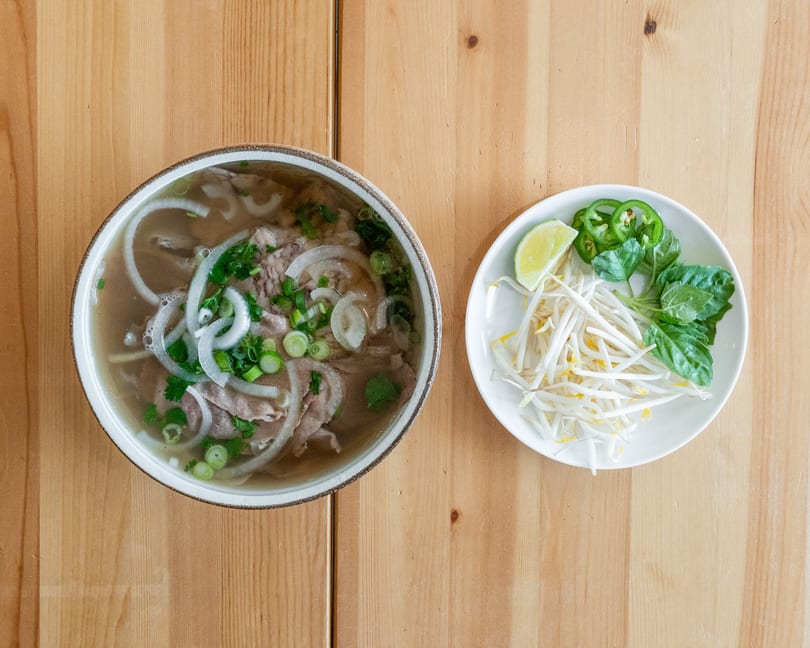 A bowl of pho with beef, noodles, and onions, alongside a plate of bean sprouts, basil, lime, and jalapeños.