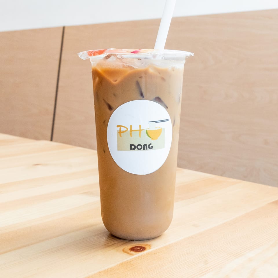 Iced coffee in a clear plastic cup with a Pho Dong logo and white straw on a wooden table.