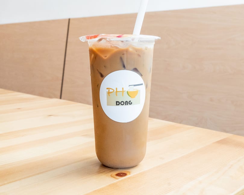 Iced coffee in a clear plastic cup with a Pho Dong logo and white straw on a wooden table.